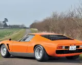 Ein leuchtend oranger Lamborghini Jota parkt auf einer einsamen Landstraße, umgeben von Feldern und kahlen Bäumen.