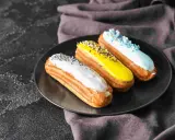 Trois éclairs gourmands sur une assiette noire : un blanc pailleté, un jaune croquant, un bleu ciel festif. La pâte à choux éclair inratable est prête à être dégustée.