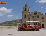 Un bus rosso "open top" per un tour di Matera, con la chiesa e i Sassi sullo sfondo.