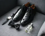 Shiny black patent leather shoes to wear with tuxedo, a bow tie, watch, and cologne are laid out on a grey couch.