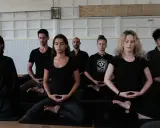 En grupp människor sitter i meditation, klädda i svarta kläder. En zen master leder sessionen.