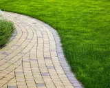 Vialetto ingresso casa curvo, pavimentato con blocchi di pietra chiara e scura, fiancheggiato da prato verde brillante e vegetazione rigogliosa.