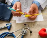 Lekarz analizuje formularz medyczny, trzymając model plemnika i długopis. Na stole leżą stetoskop, ciśnieniomierz, młoteczek neurologiczny i leki, sugerując dyskusję o skutkach ubocznych wazektomii.