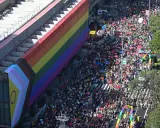 A massive crowd fills the street for what appears to be the biggest pride parade, with a giant rainbow flag draped over a building.