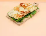 Trzy grillowane plastry sera halloumi, z czego jest, ułożone na desce z rukolą.