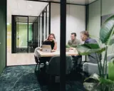 Tre kollegor skrattar vid ett mötesbord i en modern, inredd verkstad. En kvinna visar något på sin laptop.