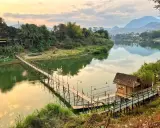 Drewniany mostek nad rzeką, bambusowa chatka i góry w tle. To jedne z wielu **laos atrakcje** warte zobaczenia.
