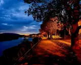 Un sentiero illuminato da lampioni lungo un fiume, con alberi dalle foglie arancioni e rosse. Una magica sera d'ottobre sui pascoli.