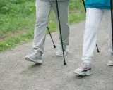 Dwie osoby maszerują z kijkami po żwirowej ścieżce, otoczone zielenią.