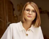 Kobieta w białej koszuli z guzikami, nosząca okulary w cienkich oprawkach, patrzy wprost.