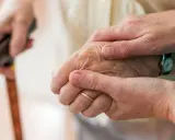 Dwie dłonie, jedna starsza, druga młodsza, trzymają się nawzajem. Laska w tle sugeruje trudności z poruszaniem się, być może związane z chorobą Parkinsona i jej dziedziczeniem.