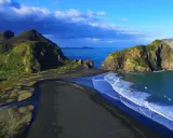 Vacker svart sandstrand med gröna kullar och klippor vid Auckland, Nya Zeeland. Havet skiftar i blått.