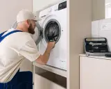 Serwisant sprawdza, czy nie ma urwanego bębna w pralce. Obok stoi walizka z narzędziami.