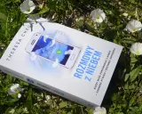 Książka "Rozmowy z niebem" leży wśród zieleni i białych kwiatów. Na okładce telefon z napisem "Niebo" i piórami.