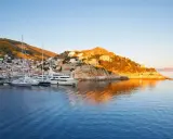 Solnedgång över hamnen på Hydra, Grekland. Lyxyachter guppar i det blå vattnet framför en vit stad som klättrar uppför en grönskande kulle.