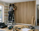Man monterar en kapa akustikpanel på väggen med en omladdningspistol. Verktyg och material ligger utspridda på golvet.