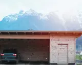 En nybyggd carport i trä med en bil parkerad under. Bakom syns snöklädda berg.