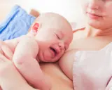Ein Baby schreit beim Stillen, die Augen geschlossen, das Gesicht verzogen. Die Mutter hält es sanft.