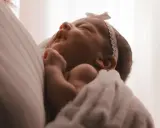Un neonato avvolto in una coperta bianca, con un fiocco rosa in testa. I suoi piccoli tremori sono quasi impercettibili mentre dorme serenamente.