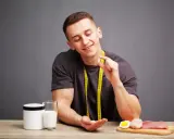 Uśmiechnięty mężczyzna trzyma tabletkę, zastanawiając się, **bcaa co to** jest. Obok stoją odżywka, mleko, jajko i mięso.