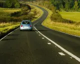 Jasnoniebieski samochód jedzie krętą drogą przez zielone pola i lasy.