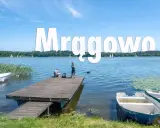 Mrągowo atrakcje: spokojny dzień nad jeziorem, dzieci łowią ryby z pomostu, żaglówki na horyzoncie.
