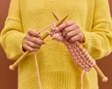 Manos tejiendo un suéter amarillo con hilo rosa, mostrando cómo hacer ochos de 4 puntos.
