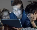 Padre y dos hijos ven una tablet, sonriendo. Podrían estar aprendiendo sobre los riesgos de las redes sociales o disfrutando de contenido familiar.