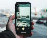 Dłoń trzyma aparat na telefon, rejestrując zimowy widok portu z zielonym statkiem i samochodami na drodze.