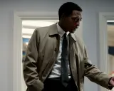 Man i trenchcoat, vit skjorta och slips, står i ett rum med fönster. Hans ansikte är i skugga, en mörk linje som framhäver hans blick.