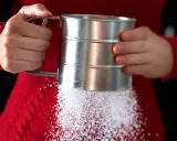 Hände halten einen Siebbecher, aus dem feiner Zucker rieselt. So einfach lässt sich puderzucker selber machen für süße Kreationen.