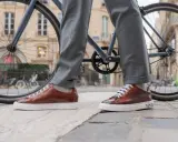 Eleganckie sneakersy do garnituru w brązowym kolorze, zestawione z szarymi spodniami, na tle miejskiego krajobrazu z rowerem.