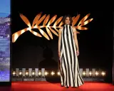A woman in a striking black and white striped gown poses on the Cannes Film Festival red carpet, bathed in the glow of the iconic Palme d'Or backdrop.