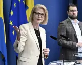 Kvinna med glasögon talar vid presskonferens med svenska och EU-flaggor bakom. Viktig information om folkräkning Sverige.