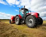 Czerwony traktor z przyczepą prasuje siano na polu. W tle błękitne niebo z chmurami. Traktorzysta w pracy.