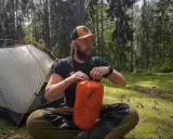 Mężczyzna w lesie pakuje pomarańczowy śpiwór, który dzięki wypełnieniu z puchu hydrofobowego ochroni go przed wilgocią.