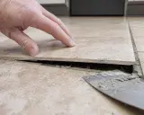 Dłoń sprawdza luźną płytkę, pod którą widać dziurę w płytce podłogowej i resztki zaprawy.