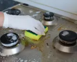 Ręka w rękawiczce czyści zabrudzoną kuchenkę gazową gąbką. Poznaj sposoby, jak wyczyścić kuchenkę, by znów lśniła.