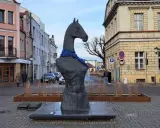 Pomnik konia w Koninie, z niebieską szarfą na szyi, stoi na brukowanym placu, otoczony kamienicami.