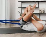 Mann liegt auf Matte, zieht Knie zur Brust und nutzt ein blaues Band zur Unterstützung. Übung zum becken mobilisieren.