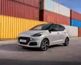 Szary Hyundai i10 N Line z czerwonymi akcentami na grillu i zderzaku, zaparkowany przed stosem kolorowych kontenerów. Sprawdź atrakcyjną hyundai i10 cena!