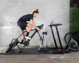 Kobieta ćwiczy na rowerze stacjonarnym, który jest rodzajem trenażera. Obok stoi biurko na laptopa i wentylator.