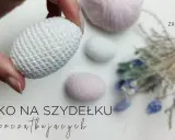 Ręcznie robione, białe jajko na szydełku, idealne na początek przygody z szydełkowaniem. W tle pastelowe włóczki i suszone kwiaty.