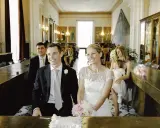Uśmiechnięty świadek na ślubie, obok panny młodej, w eleganckim garniturze. W tle goście i dekoracje sali.