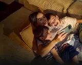 A mother and two sons laugh while looking at a smartphone on a couch.