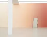A gallery wall features a stunning gradient in art, transitioning from soft peach to warm coral. A rough stone slab leans against it.