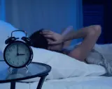 Osoba w łóżku zakrywa uszy, budzik wskazuje 3:00. To obraz bezsenności i zaburzeń snu.