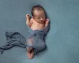 Neonato dorme avvolto in un telo blu, con le braccia sollevate e i pugni chiusi.