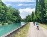 Bambini in bicicletta lungo la ciclovia del Mincio, con il canale turchese e filari di cipressi.
