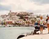 A gay couple walks hand-in-hand along a harbor in Spain, with a white-washed town and ancient fortress rising behind them.
